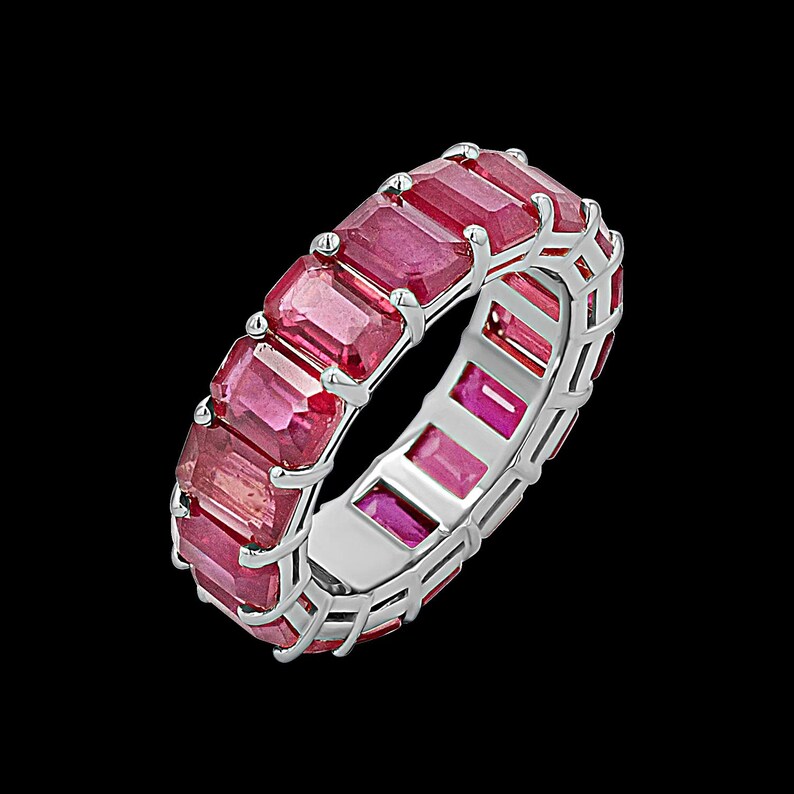 May include: A silver-toned eternity band ring featuring a continuous row of rectangular, emerald-cut red gemstones set in a prong setting. The ring has a polished metallic finish and showcases a series of vibrant, deep pinkish-red stones.