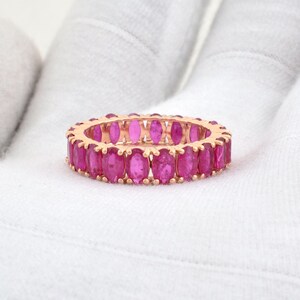 Può includere: Un anello dorato con una fila continua di pietre preziose rosa vibranti a taglio ovale. Il design dell'anello presenta uno stile a fascia eternity completa, con le pietre preziose molto vicine tra loro. L'anello è esposto su una superficie bianca.