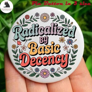 May include: A white pin button with the text "Radicalized by Basic Decency" in a retro font, surrounded by a floral wreath. The pin has a glossy finish and is likely a wearable accessory.
