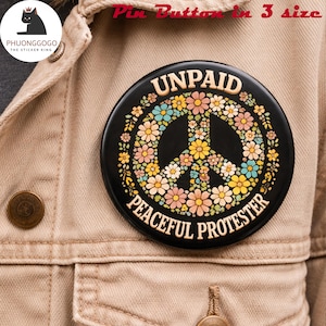 Puede incluir: Un botón de pin negro con las palabras "UNPAID PEACEFUL PROTESTER" que rodean un símbolo de la paz hecho de flores coloridas. El pin es redondo y está sujeto a una chaqueta beige.