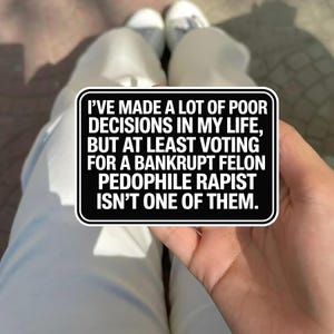 May include: A black and white sticker with the text: "I'VE MADE A LOT OF POOR DECISIONS IN MY LIFE, BUT AT LEAST VOTING FOR A BANKRUPT FELON PEDOPHILE RAPIST ISN'T ONE OF THEM." The sticker is held by a person.