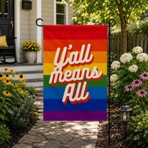 May include: A rectangular garden flag featuring a horizontal rainbow stripe pattern in red, orange, yellow, green, blue, and purple. The text "Y'all means All" is printed in a large, white, cursive font with a red drop shadow across the centre.