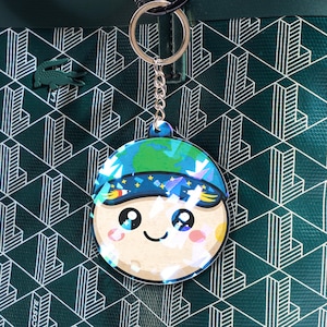 May include: A round keychain featuring a cartoon character with a blue and green Earth-themed hat and a smiling face. The keychain hangs from a silver chain and ring. The background is a dark green tote bag with a geometric pattern and a crocodile logo.