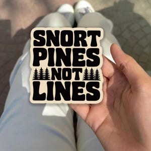 May include: A beige sticker with the text "SNORT PINES NOT LINES" in a bold, black font. The word "NOT" is separated by a row of stylized pine trees. The sticker is held in a hand.