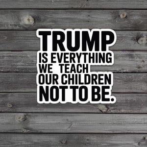 Puede incluir: Una pegatina blanca con texto negro que dice "TRUMP IS EVERYTHING WE TEACH OUR CHILDREN NOT TO BE." La pegatina está sobre un fondo de madera gris desgastada.