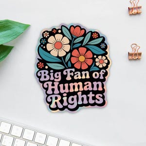 May include: A holographic sticker with a floral design and the text "Big Fan of Human Rights." The sticker features flowers in shades of orange, cream, and blue, with green leaves. The text is in a retro font.