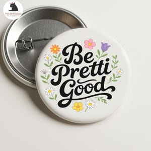 Puede incluir: Un pin de botón blanco con la frase "Be Pretti Good" en escritura negra, rodeada de flores coloridas y hojas verdes. El pin tiene una parte trasera de metal con un cierre de imperdible. Un accesorio alegre.