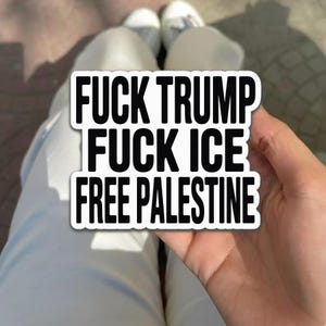 May include: A white sticker with black text that reads "FUCK TRUMP FUCK ICE FREE PALESTINE". The sticker is held in a hand, with white pants and shoes visible in the background.