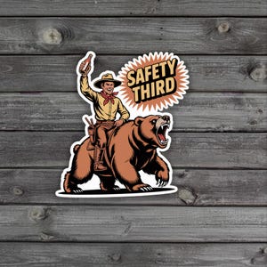 May include: A sticker featuring a cowboy riding a brown bear, holding a bottle aloft. The cowboy wears a hat, yellow shirt, and red neckerchief. The text "SAFETY THIRD" is in a burst shape.