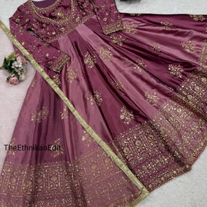 May include: A long, flowing, plum-colored dress with intricate gold embroidery. The dress features long sleeves and a round neckline, with detailed patterns on the bodice and skirt. A gold trim and a matching necklace complete the elegant look. The text "TheEthnikaaEdit" is visible.
