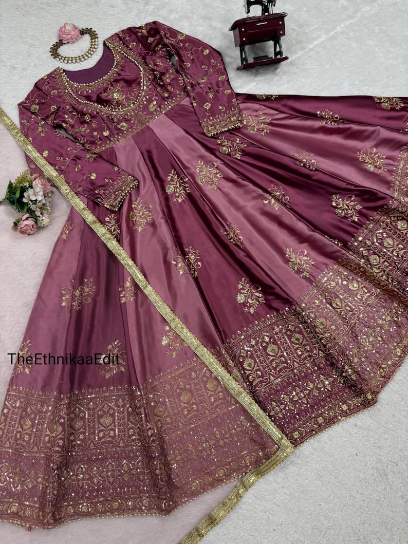May include: A long, flowing, plum-colored dress with intricate gold embroidery. The dress features long sleeves and a round neckline, with detailed patterns on the bodice and skirt. A gold trim and a matching necklace complete the elegant look. The text "TheEthnikaaEdit" is visible.