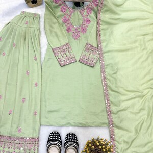 May include: A light green embroidered salwar kameez set with a matching dupatta. The set features intricate pink embroidery on the neckline, sleeves, and hem. A pair of black and white beaded flats are at the bottom of the image.