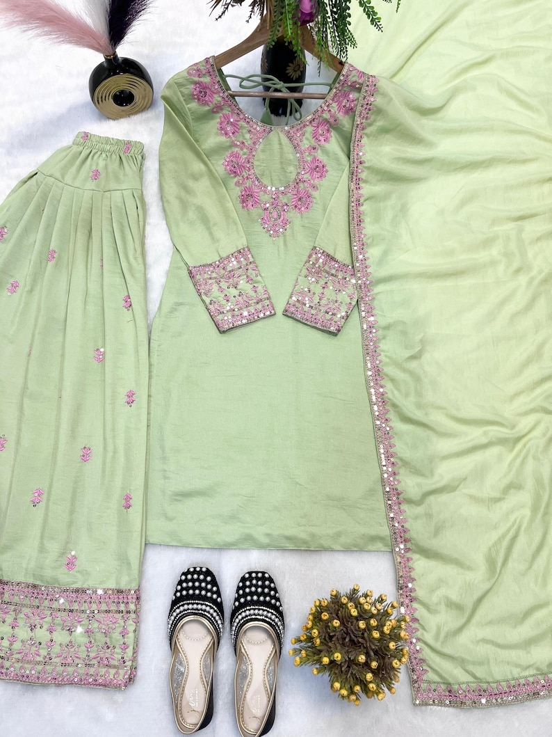 May include: A light green embroidered salwar kameez set with a matching dupatta. The set features intricate pink embroidery on the neckline, sleeves, and hem. A pair of black and white beaded flats are at the bottom of the image.