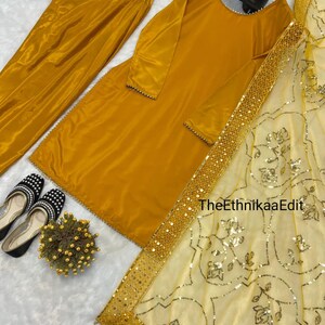 May include: Mustard yellow women's suit set, including a long-sleeved tunic, matching pants, and a sheer scarf with gold embellishments. The tunic and pants are made of a shiny fabric. Black and white shoes with a floral accent are also visible.