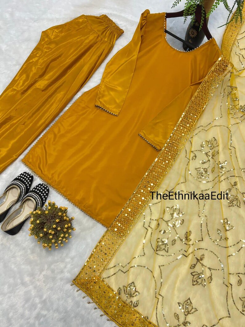May include: Mustard yellow women's suit set, including a long-sleeved tunic, matching pants, and a sheer scarf with gold embellishments. The tunic and pants are made of a shiny fabric. Black and white shoes with a floral accent are also visible.