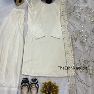 May include: Cream-colored salwar kameez set with matching pants and a dupatta. The long-sleeved tunic has gold trim. Black and gold embellished flats and a small floral arrangement complete the ensemble. TheEthnikaaEdit is printed on the image.