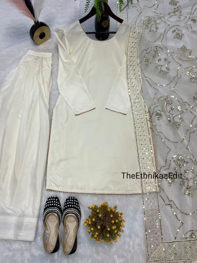 May include: Cream-colored salwar kameez set with matching pants and a dupatta. The long-sleeved tunic has gold trim. Black and gold embellished flats and a small floral arrangement complete the ensemble. TheEthnikaaEdit is printed on the image.