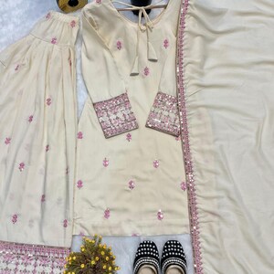 May include: Cream-colored three-piece ethnic wear set with pink floral embroidery and sequin detailing. The set includes a long-sleeved tunic, wide-leg pants, and a matching scarf. A pair of black and white beaded flats with gold trim complete the ensemble.