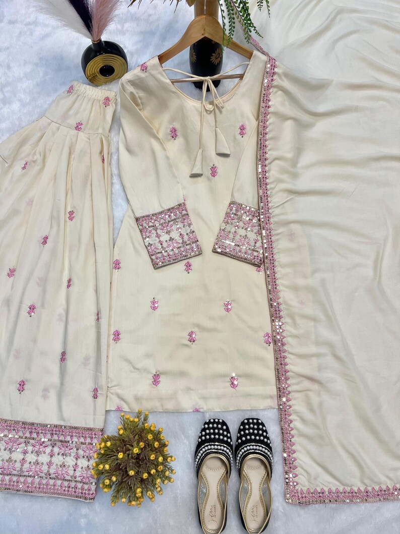 May include: Cream-colored three-piece ethnic wear set with pink floral embroidery and sequin detailing. The set includes a long-sleeved tunic, wide-leg pants, and a matching scarf. A pair of black and white beaded flats with gold trim complete the ensemble.