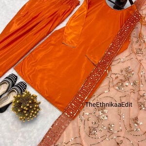 May include: An orange satin salwar kameez set. The set includes a long tunic, matching pants, and a sheer peach dupatta with gold sequin embellishments. The dupatta has a decorative border. Black and white shoes are visible in the corner.