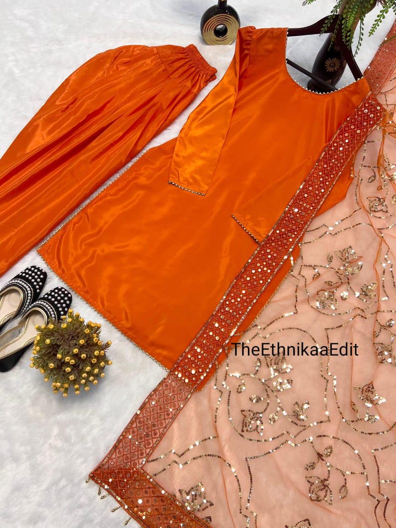 May include: An orange satin salwar kameez set. The set includes a long tunic, matching pants, and a sheer peach dupatta with gold sequin embellishments. The dupatta has a decorative border. Black and white shoes are visible in the corner.