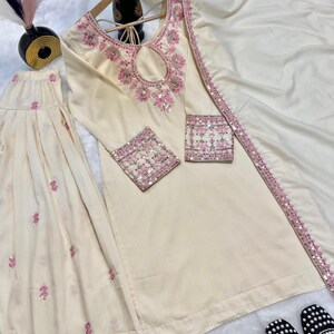 May include: Cream-colored embroidered kurta suit with a matching dupatta and palazzo pants. The kurta features pink floral embroidery around the neckline and cuffs. The dupatta has a pink sequined border. Black and white embroidered flats are at the bottom.