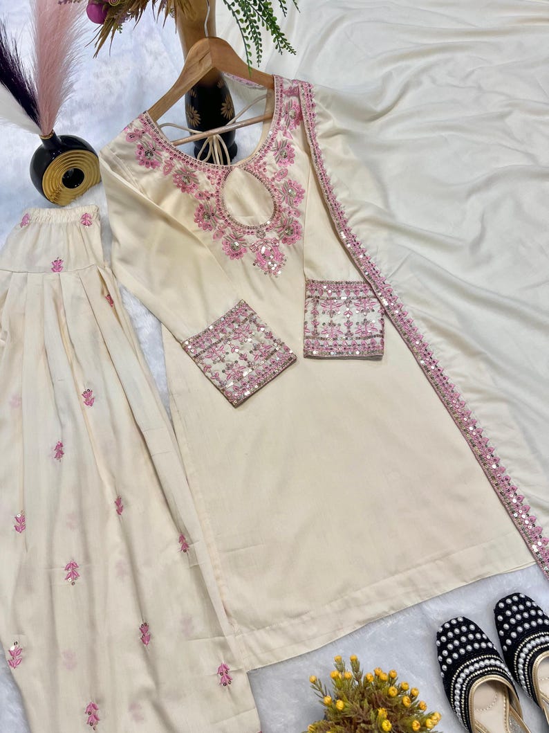 May include: Cream-colored embroidered kurta suit with a matching dupatta and palazzo pants. The kurta features pink floral embroidery around the neckline and cuffs. The dupatta has a pink sequined border. Black and white embroidered flats are at the bottom.