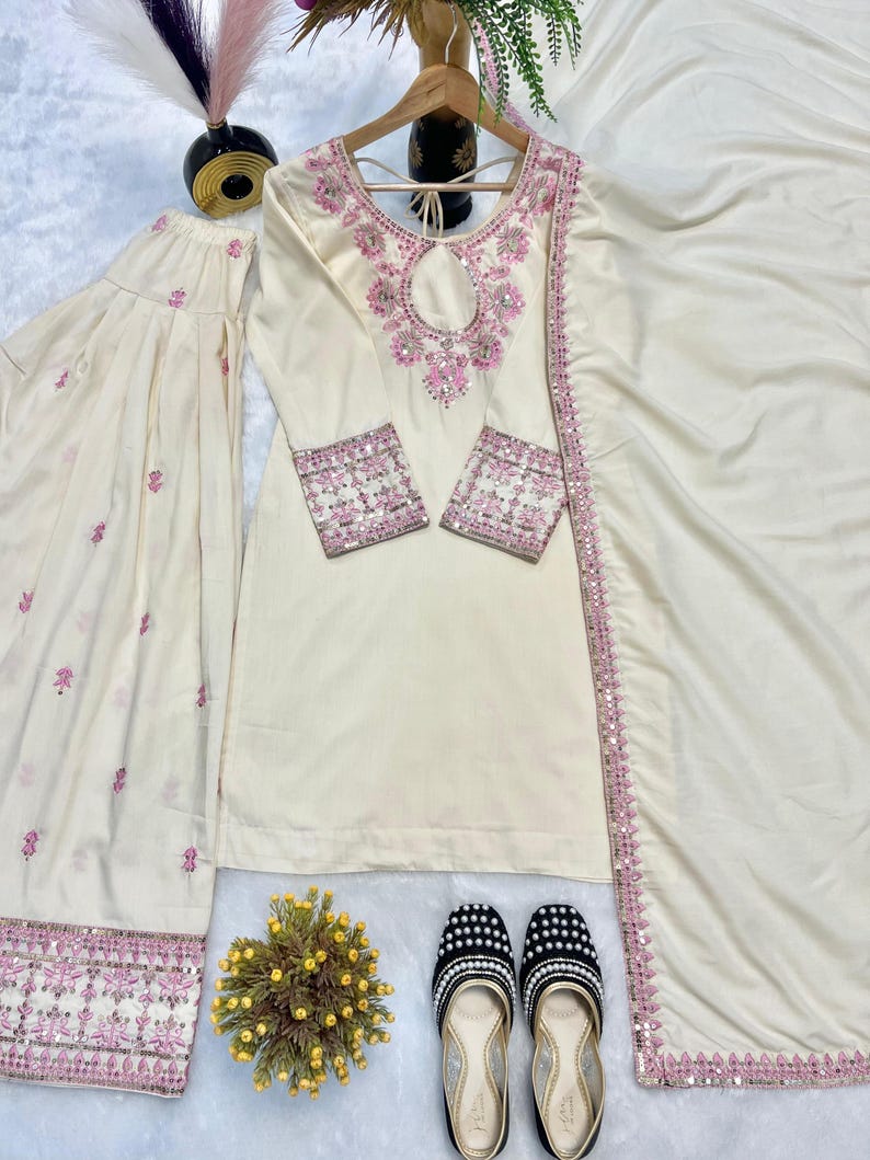 May include: Cream-colored embroidered Indian suit set with a matching dupatta and pants. The suit features pink floral embroidery on the neckline, sleeves, and pants. Black and white beaded flats and a small yellow floral arrangement complete the ensemble.