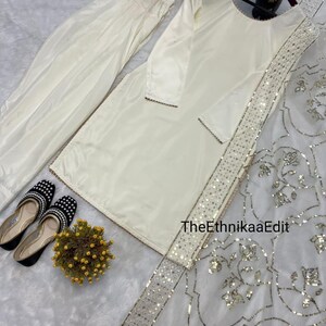 May include: Cream-colored traditional outfit, including a long-sleeved tunic, pants, and a sheer scarf with silver embellishments. The outfit is paired with black and white beaded shoes and a small floral arrangement. TheEthnikaaEdit is visible on the scarf.