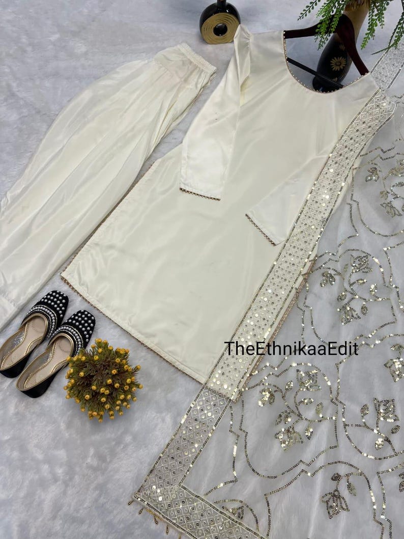 May include: Cream-colored traditional outfit, including a long-sleeved tunic, pants, and a sheer scarf with silver embellishments. The outfit is paired with black and white beaded shoes and a small floral arrangement. TheEthnikaaEdit is visible on the scarf.