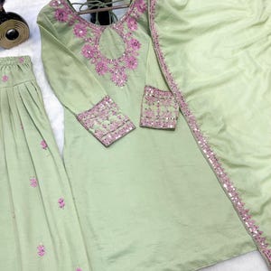 May include: A light green, three-piece embroidered outfit. The long-sleeved top and skirt are adorned with pink floral embroidery and sequins. A matching scarf is draped over the outfit. The outfit is displayed on a wooden hanger.