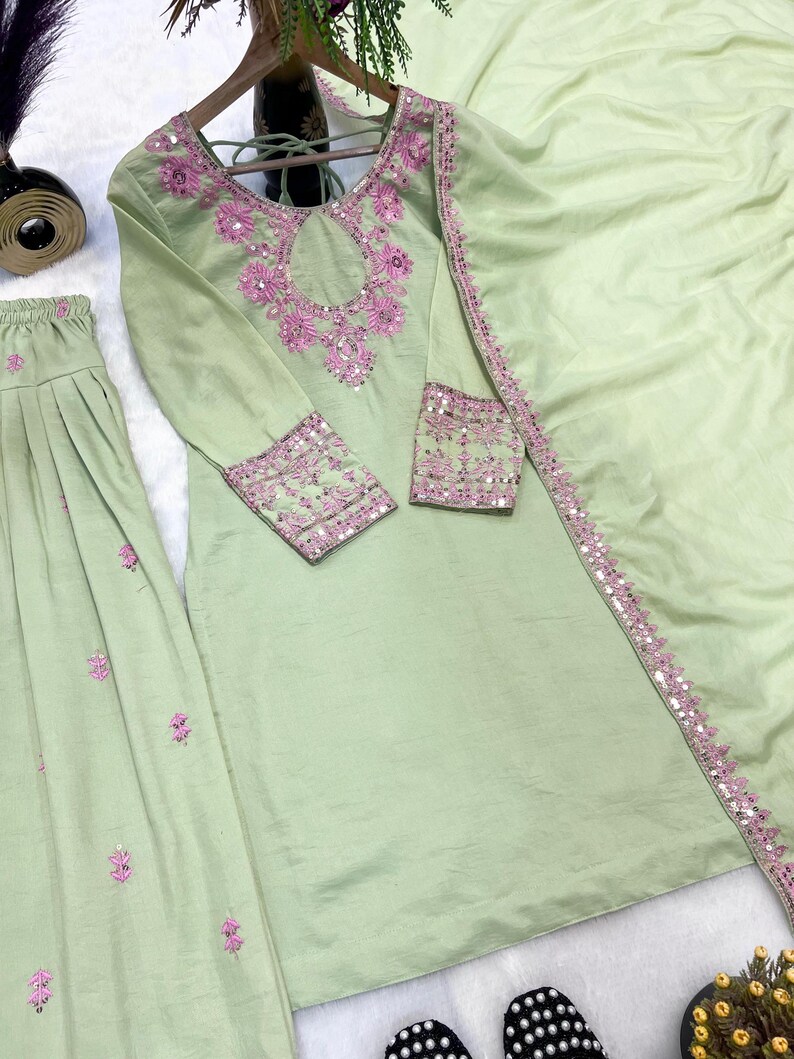 May include: A light green, three-piece embroidered outfit. The long-sleeved top and skirt are adorned with pink floral embroidery and sequins. A matching scarf is draped over the outfit. The outfit is displayed on a wooden hanger.