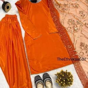May include: An orange satin salwar kameez set with matching pants and a peach-colored dupatta. The outfit is accessorized with black and white beaded flats. The text "TheEthnikaaEdit" is visible on the right side of the image.