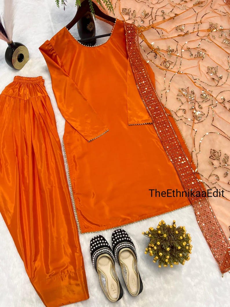 May include: An orange satin salwar kameez set with matching pants and a peach-colored dupatta. The outfit is accessorized with black and white beaded flats. The text "TheEthnikaaEdit" is visible on the right side of the image.