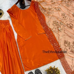 May include: An orange salwar kameez set with a matching dupatta. The long-sleeved top and pants are made of a shiny fabric. The dupatta is peach with gold embellishments and a sequined border. Black and white embroidered shoes are at the bottom.