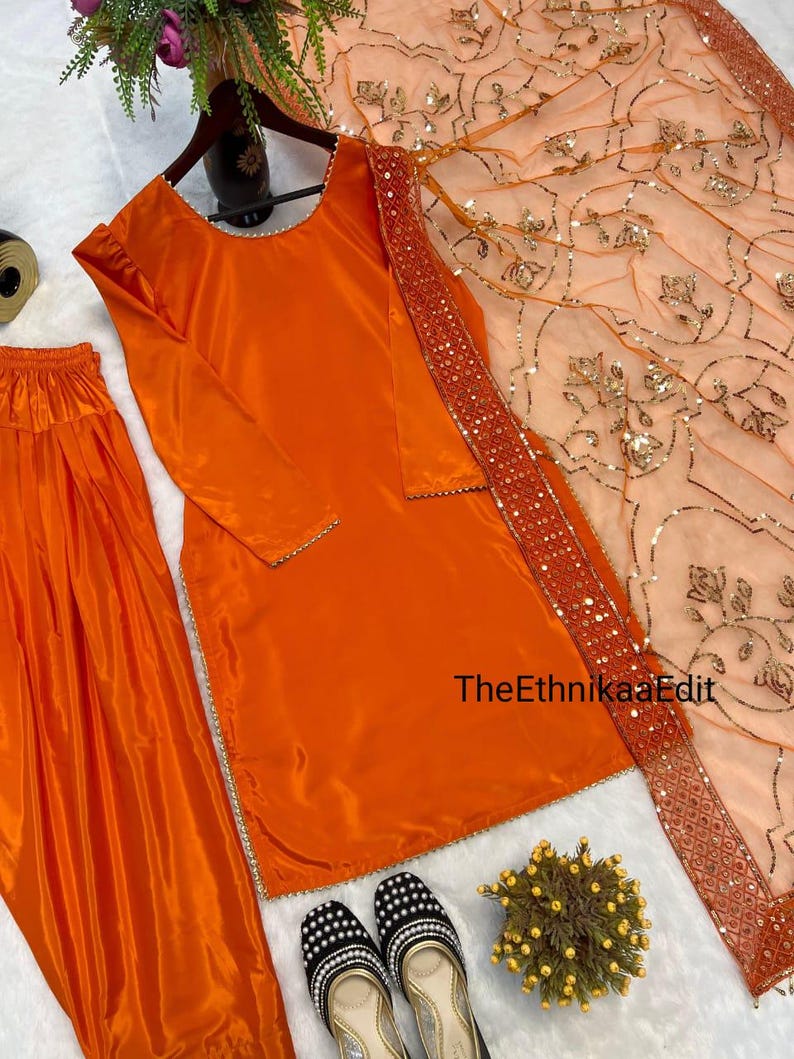 May include: An orange salwar kameez set with a matching dupatta. The long-sleeved top and pants are made of a shiny fabric. The dupatta is peach with gold embellishments and a sequined border. Black and white embroidered shoes are at the bottom.