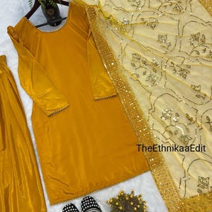May include: Mustard yellow salwar kameez set with a matching dupatta. The long-sleeved tunic and pants are made of a shiny fabric. The dupatta is light yellow with floral embroidery and a gold border. Black and white embroidered shoes are at the bottom.