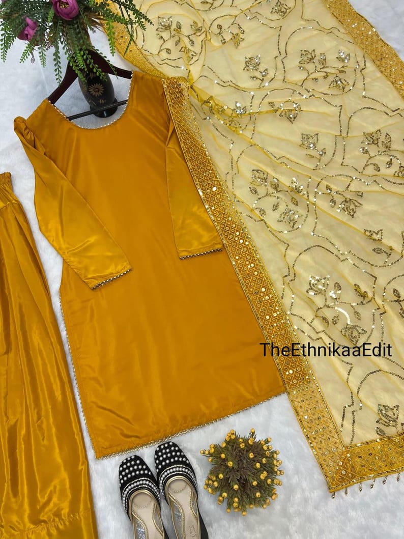 May include: Mustard yellow salwar kameez set with a matching dupatta. The long-sleeved tunic and pants are made of a shiny fabric. The dupatta is light yellow with floral embroidery and a gold border. Black and white embroidered shoes are at the bottom.
