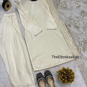 May include: Ivory-colored satin salwar kameez set with matching pants and a dupatta. The outfit features gold trim and is paired with black and gold embellished flats. The EthnikaaEdit text is visible on the dupatta.