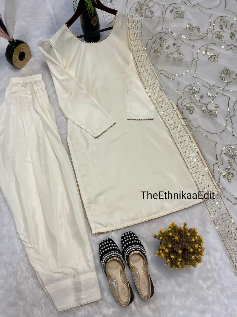 May include: Ivory-colored satin salwar kameez set with matching pants and a dupatta. The outfit features gold trim and is paired with black and gold embellished flats. The EthnikaaEdit text is visible on the dupatta.