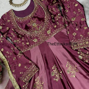 May include: A formal, floor-length burgundy dress with long sleeves and intricate gold embroidery. The dress features a round neckline and a fitted bodice that flows into a pleated skirt. A matching necklace and floral hair accessory are also visible.