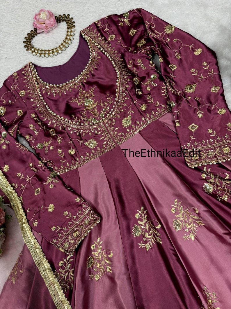 May include: A formal, floor-length burgundy dress with long sleeves and intricate gold embroidery. The dress features a round neckline and a fitted bodice that flows into a pleated skirt. A matching necklace and floral hair accessory are also visible.