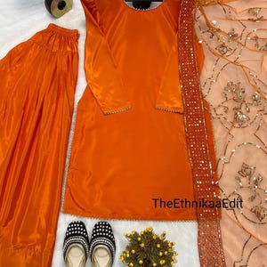 May include: An orange salwar kameez set with a matching dupatta. The set includes a long-sleeved tunic, loose trousers, and a sheer scarf with sequin trim. The outfit is paired with black and silver embellished flats.