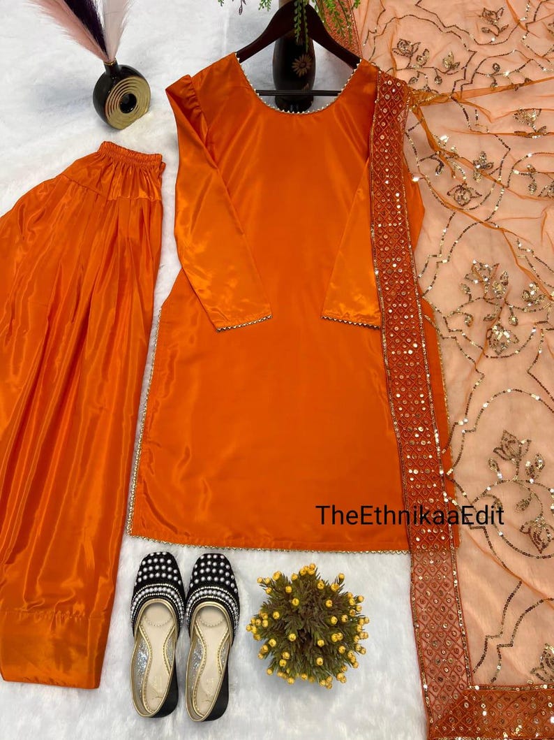May include: An orange salwar kameez set with a matching dupatta. The set includes a long-sleeved tunic, loose trousers, and a sheer scarf with sequin trim. The outfit is paired with black and silver embellished flats.