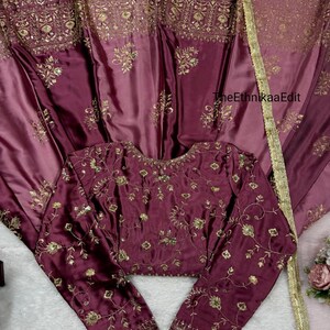 May include: A burgundy-colored, embroidered blouse with long sleeves and a matching skirt. The set features intricate gold floral embroidery and sequin detailing. The fabric has a satin sheen, and the skirt has a gold trim. The text "TheEthnikaaEdit" is visible.