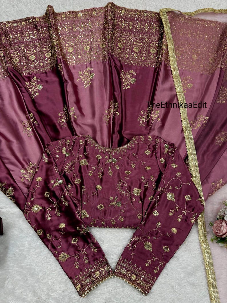 May include: A burgundy-colored, embroidered blouse with long sleeves and a matching skirt. The set features intricate gold floral embroidery and sequin detailing. The fabric has a satin sheen, and the skirt has a gold trim. The text "TheEthnikaaEdit" is visible.