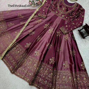 May include: A burgundy satin gown with intricate gold embroidery. The long-sleeved dress features a fitted bodice and a flowing skirt. The EthnikaaEdit text is visible on the image. A matching scarf with gold trim is draped over the dress.