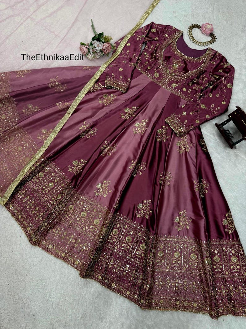 May include: A burgundy satin gown with intricate gold embroidery. The long-sleeved dress features a fitted bodice and a flowing skirt. The EthnikaaEdit text is visible on the image. A matching scarf with gold trim is draped over the dress.