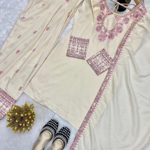 May include: Cream-colored embroidered kurta suit with matching pants and dupatta. The kurta features pink floral embroidery and sequin detailing. Black and white beaded flats are also included.