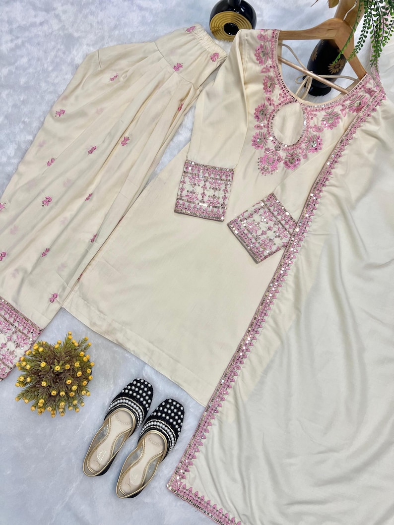 May include: Cream-colored embroidered kurta suit with matching pants and dupatta. The kurta features pink floral embroidery and sequin detailing. Black and white beaded flats are also included.